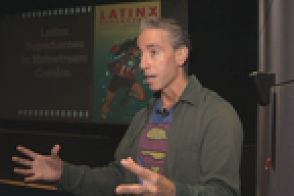 Prof. Aldama speaking in a "Superman" t-shirt. The word "Latinx" and an image of a female superhero are visible behind him. 